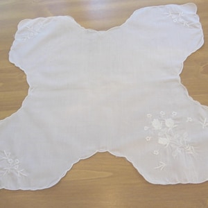 May include: A white, embroidered bib-shaped cloth with a scalloped edge. Delicate floral embroidery accents the corners and edges of the cloth. The cloth is made of a thin, semi-transparent fabric.