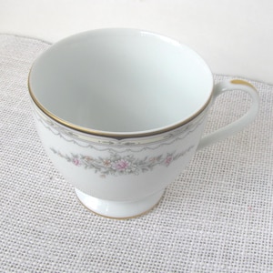 May include: White teacup with a gold rim and handle. The cup features a floral design with pink and gray accents. The teacup is on a white pedestal base and is likely made of porcelain.