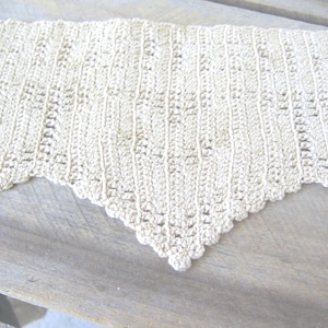 May include: Cream-colored, handmade crochet shawl with a scalloped edge. The shawl features a detailed openwork pattern and a triangular shape. The texture of the yarn is visible, suggesting a soft, natural fiber. The shawl is displayed on a wooden surface.