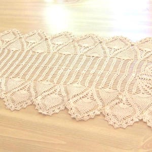 May include: A cream-colored, hand-crocheted table runner with a detailed, openwork design. The runner features a central, ribbed section flanked by decorative, scalloped edges. The intricate pattern adds a touch of vintage charm to any table setting.