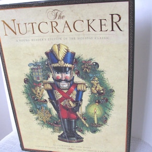 May include: A book cover for "The Nutcracker" features a detailed illustration of a nutcracker soldier in a blue and red uniform, surrounded by a Christmas wreath. The title is in elegant gold lettering, with additional text indicating it's a young reader's edition.