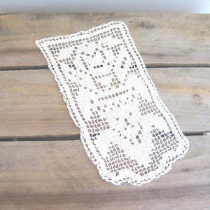 May include: An off-white, rectangular doily with a delicate, open-weave crochet design. The edges are scalloped, adding a decorative touch. The doily is placed on a weathered, light brown wooden surface, creating a vintage aesthetic.