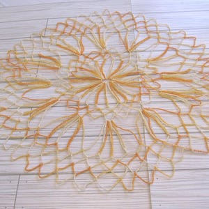 May include: A handmade, circular doily with an openwork design. The doily features a central floral pattern in shades of yellow and orange, with a delicate, web-like border. The piece is placed on a light-colored wooden surface.