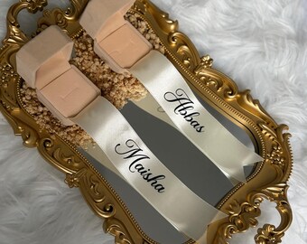 May include: Two beige velvet ring boxes are placed on a gold ornate mirror tray. Each box has a white satin ribbon attached, printed with the names Maisha and Abbas in black script. The tray is decorated with small dried flowers.