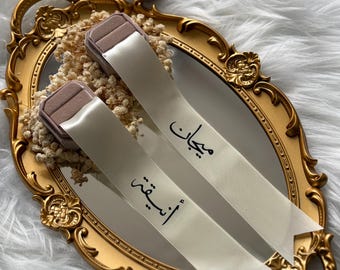 May include: A gold-framed mirror with a decorative edge holds two ring boxes and two ribbons. The ribbons are cream-colored with black Arabic script. The ring boxes are a muted mauve color.