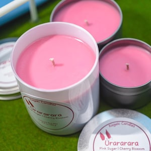 May include: A selection of pink soy wax candles in white and silver containers. The candles have wicks and are labelled "Urararara Pink Sugar | Cherry Blossom". The candles are on a green surface.