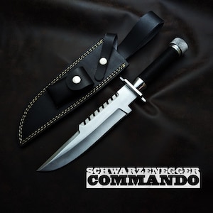 May include: A large, silver and black survival knife with a serrated blade, resting next to its black leather sheath. The knife has a textured silver handle and a black grip. The text "SCHWARZENEGGER COMMANDO" is printed in white.