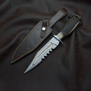 Może przedstawiać: Ręcznie wykonany nóż myśliwski z ząbkowanym ostrzem i rękojeścią z poroża jelenia, obok skórzanej pochwy. Ostrze ma inskrypcję. Pochwa jest brązowa z ozdobnymi przeszyciami i zapięciem na guzik. Nóż leży na ciemnobrązowej powierzchni.