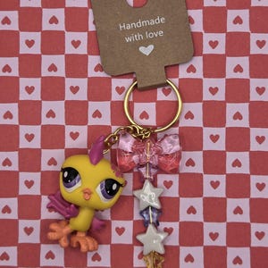 Puede incluir: Un llavero hecho a mano con una figura de pájaro amarillo con ojos morados y alas rosas. El llavero incluye un lazo rosa, cuentas en forma de estrella y un dije de llave dorada. La etiqueta dice "Handmade with love."