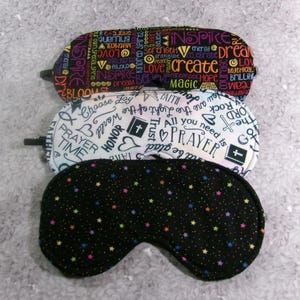 May include: Three sleep masks with different patterns. The top mask has a black background with colorful words like "inspire" and "create." The middle mask is white with religious symbols and words. The bottom mask is black with colorful stars.
