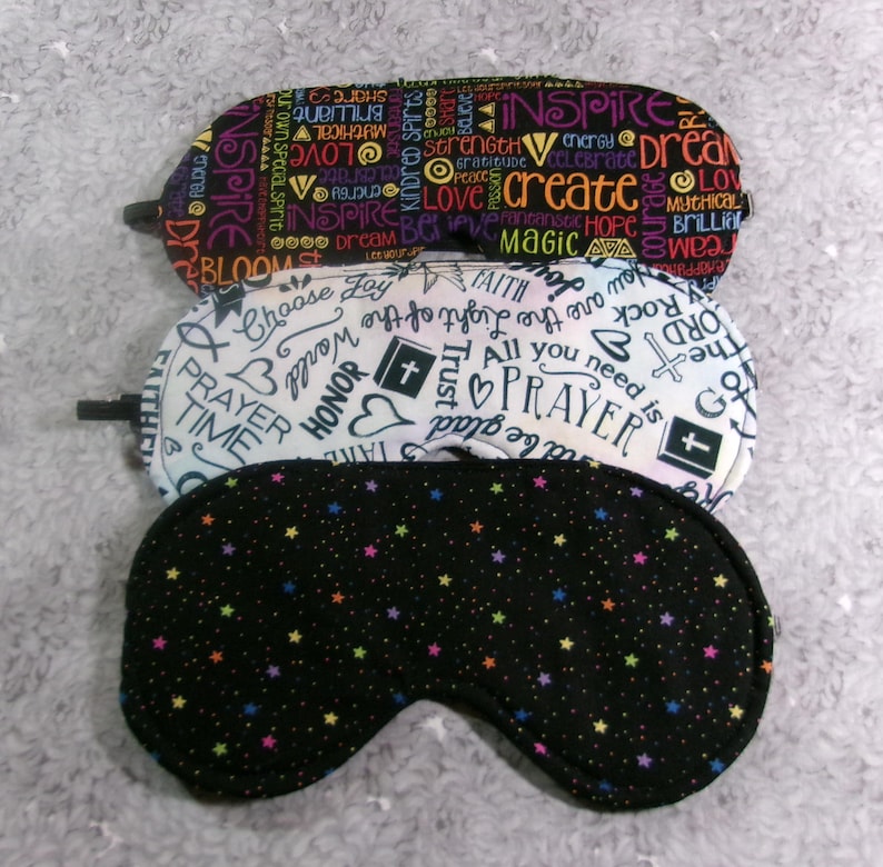 May include: Three sleep masks with different patterns. The top mask has a black background with colorful words like "inspire" and "create." The middle mask is white with religious symbols and words. The bottom mask is black with colorful stars.