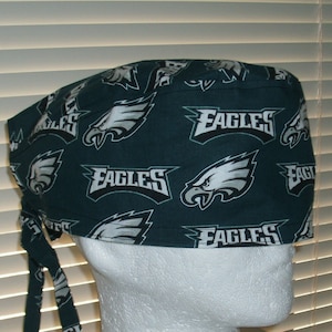 May include: Green surgical scrub hat with a white and gray repeated pattern of the Philadelphia Eagles logo and the word "Eagles".