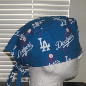 May include: Blue and white surgical scrub hat with a Los Angeles Dodgers pattern. The hat has a tie on the back and features the team's logo and the words "Dodgers" and "LA".