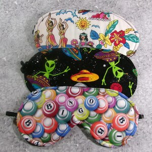 May include: Three sleep masks with different patterns. One features hula dancers, another has aliens and UFOs against a starry background, and the third displays bingo balls with the word "BINGO". Each mask has a black elastic strap.