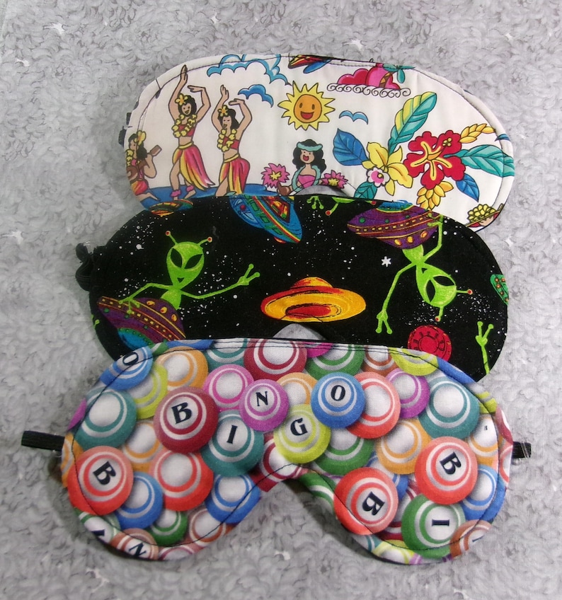 May include: Three sleep masks with different patterns. One features hula dancers, another has aliens and UFOs against a starry background, and the third displays bingo balls with the word "BINGO". Each mask has a black elastic strap.