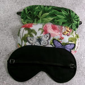 May include: Three sleep masks with elastic straps. The top mask has a green cannabis leaf pattern. The middle mask features butterflies and roses on a light blue background. The bottom mask is solid black.