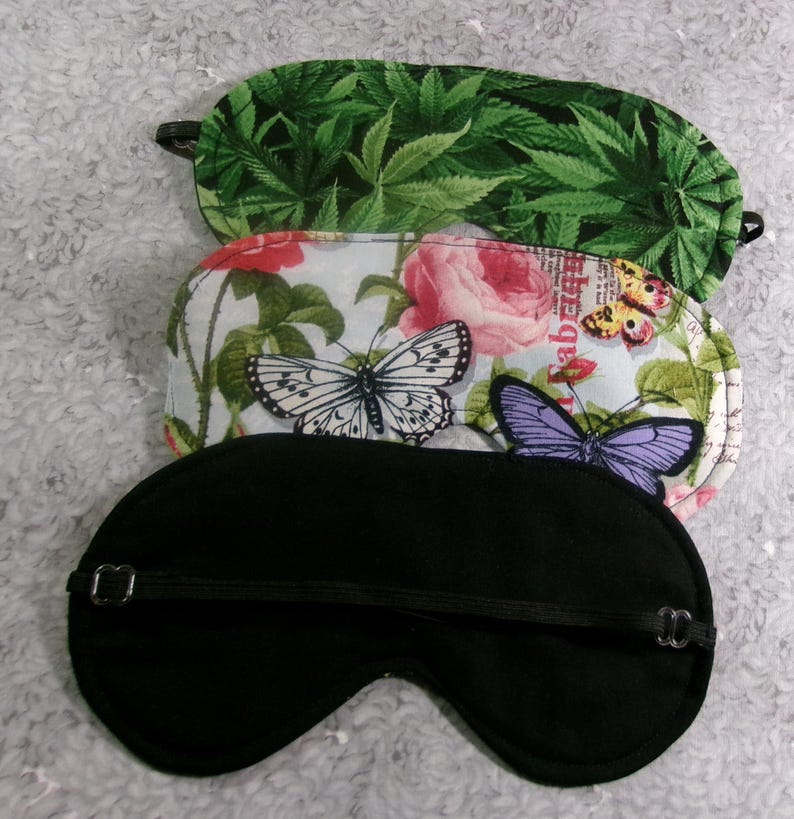 May include: Three sleep masks with elastic straps. The top mask has a green cannabis leaf pattern. The middle mask features butterflies and roses on a light blue background. The bottom mask is solid black.