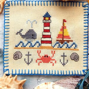 Op de afbeelding: Een vierkant kruissteekwerk op crèmekleurige stof met een blauwe festonsteekrand. Het ontwerp toont een rood-witte vuurtoren, een grijze walvis, een zeilboot, een rode krab, twee ankers en twee schelpen, geborduurd met gekleurde draad.