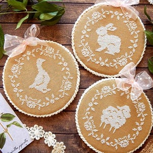 Könnte beinhalten: Drei runde Stickereien mit weißen Tiersilhouetten: ein Hase, ein Schaf und eine Ente, jeweils umgeben von einem floralen Kranz. Sie sind mit rosa Bändern gebunden. Der Hintergrund ist eine Holzoberfläche.