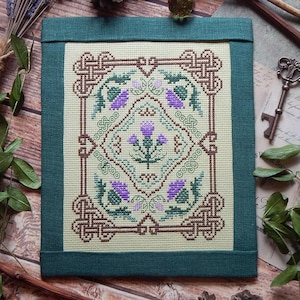 Può includere: Un ricamo a punto croce incorniciato, con un motivo centrale di cardo nei toni del viola e del verde, circondato da intricati nodi. L'opera è ambientata su uno sfondo color crema e incorniciata con tessuto verde scuro. Elementi decorativi includono una chiave e rametti di lavanda.