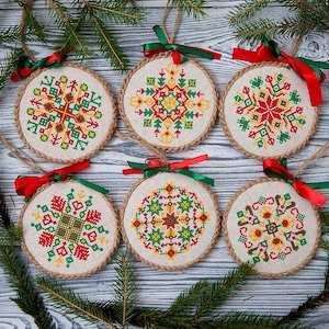 Puede incluir: Seis adornos navideños redondos con patrones geométricos bordados en rojo, verde y amarillo. Cada adorno tiene un borde de cuerda de yute y está atado con una cinta roja y verde. Dispuestos sobre una superficie de madera con ramas de pino.