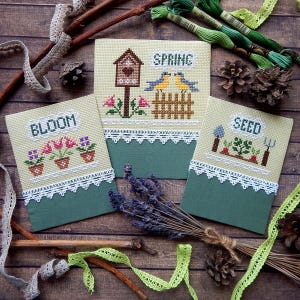 May include: Three cross-stitch panels with spring designs. One says "BLOOM" with potted flowers, another says "SPRING" with birds and a birdhouse, and the third says "SEED" with garden tools and vegetables. Each panel has a green base and white lace trim.