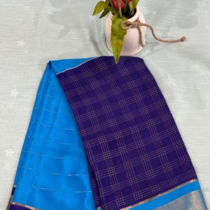 May include: A folded saree featuring a vibrant blue section with horizontal silver lines and a deep purple section with a gold grid pattern. The fabric is accented by a wide border with intricate gold metallic detailing along the bottom edge.