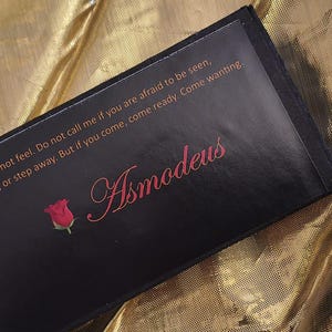 May include: A black rectangular box with the word "Asmodeus" in pink script, a small red rose, and text that reads "Pretend you do not feel. Do not call me if you are afraid to be seen, Step forward, or step away. But if you come, come ready. Come wanting."