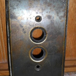 May include: A rectangular, antique brass light switch plate with three circular holes. The plate has a dark, aged patina with signs of wear and tear, and a gold-colored border.
