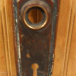 May include: Antique bronze door plate with a keyhole and a circular opening. The plate has a decorative, geometric design and a distressed finish, showing signs of age and wear. The plate is mounted on a wooden surface.