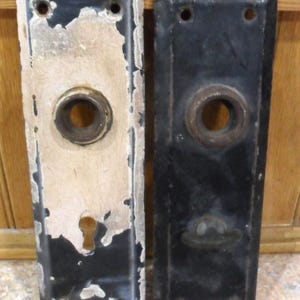 May include: Two antique door plates. One plate has black paint with chipped areas revealing a beige undercoat. The other plate is black with a circular opening and a decorative knob. Both plates have screw holes.