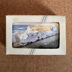 May include: A rectangular box with a framed image of a passenger train traveling along a coastal track. The train is silver, blue, and red, with a white front. The background shows a coastline with cliffs and ocean.