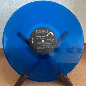May include: A blue vinyl record of Elvis Presley's "Moody Blue" album, displayed on a wooden stand. The record label is visible, with the RCA logo and song titles. The record is a vibrant shade of blue.