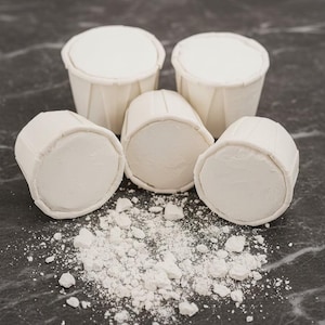 May include: Five white, cylindrical chalk blocks with pleated paper wrappers, arranged in a cluster. The chalk pieces have a smooth, flat top surface, and a small pile of white chalk dust is scattered in front of the group on the surface.