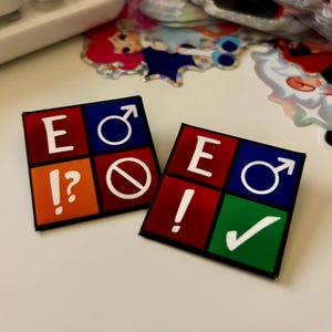 May include: Two square pins with a black border. Each pin has four colored squares: red, orange, blue, and green. The pins feature the letter "E", an exclamation point, a question mark, a male symbol, a no symbol, and a check mark.