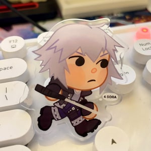 May include: A clear acrylic charm featuring a cartoon character with spiky white hair, black jacket, and holding a black weapon. The character is running with a determined expression. The text "4 SORA" is visible.