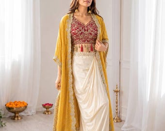 May include: A three-piece ensemble featuring a red embroidered crop top, a cream-colored draped skirt, and a yellow patterned cape with gold trim and red tassels. The outfit is designed for a formal occasion.