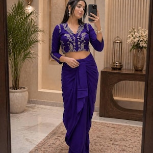 May include: A woman in a two-piece purple outfit. The top is a long-sleeved crop top with detailed embroidery around the neckline and sleeves. The bottom is a draped skirt. The outfit is a vivid purple colour.