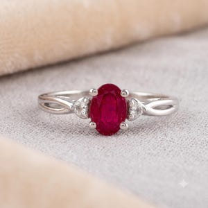 Peut inclure: Une bague argentée ornée d'un rubis rouge foncé taille ovale maintenu par quatre griffes. L'anneau se divise en deux brins délicats de chaque côté, avec une petite pierre d'accent ronde et transparente sertie là où les brins se rejoignent.