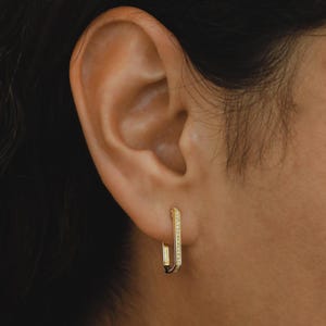 Könnte beinhalten: Goldfarbene Creolen mit rechteckigem Design. Die Ohrringe haben eine Reihe kleiner, funkelnder Steine entlang der Vorderkante. Die Ohrringe sind an einem Ohr abgebildet.