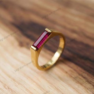 May include: A gold ring featuring a single rectangular baguette-cut deep red gemstone set horizontally in the center. The band is smooth and polished, with a solid metal construction that frames the vibrant red stone securely in a bezel setting.