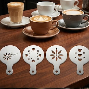Pode incluir: Estênceis de café em aço inoxidável com vários desenhos, incluindo uma flor, corações e uma estrela. Várias xícaras de café com arte latte são visíveis em uma superfície de madeira, junto com uma máquina de espresso profissional.