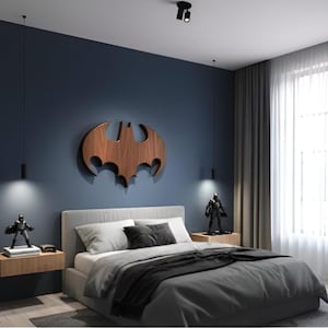 May include: A bedroom with a dark blue wall featuring a wooden Batman logo. The bed has gray bedding and pillows. Two small Batman figurines are on the nightstands. The room has a modern design with hanging lights and a window with sheer curtains.