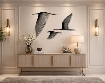 May include: Three black metal bird wall art pieces in flight are mounted on a cream-colored wall. A beige cabinet with gold accents and a lamp sit below the artwork. A vase of white flowers and a potted tree are also in the room.