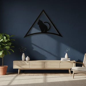 Op de afbeelding: Zwart metalen katten silhouet in een driehoekig frame, gemonteerd op een donkerblauwe muur. De kamer heeft een houten kast, een plant in een pot en een rieten stoel, wat een moderne, minimalistische esthetiek creëert.