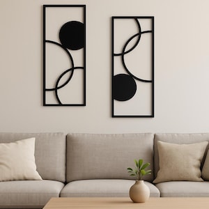 Puede incluir: Dos piezas de arte mural de metal negro con diseños geométricos. Cada pieza presenta un marco rectangular con círculos superpuestos y un círculo sólido. El arte está montado en una pared de color neutro sobre un sofá beige.
