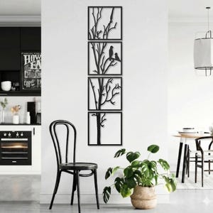 May include: Four black metal wall art panels depicting tree branches and birds. The panels are square and arranged vertically. The art is displayed on a white wall, with a black chair and a potted plant in the foreground.