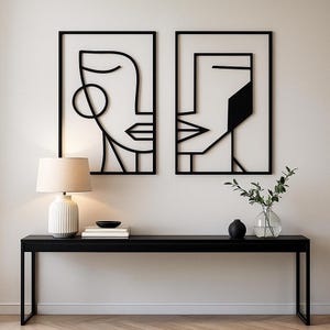 Puede incluir: Dos obras de arte murales enmarcadas en negro con diseños de rostros abstractos cuelgan sobre una consola negra. Una lámpara blanca con pantalla beige, un cuenco negro y objetos decorativos están sobre la mesa. La pared está pintada de blanco.