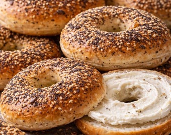 May include: A close-up shot of several bagels, some whole and others sliced open. The bagels are golden brown, covered in sesame seeds and poppy seeds. One bagel is cut open and spread with a thick layer of cream cheese, ready to eat.