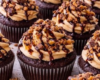 May include: A close-up shot of several chocolate cupcakes with peanut butter frosting, drizzled with chocolate sauce, and topped with chopped peanut butter cups. The cupcakes are in brown paper liners.